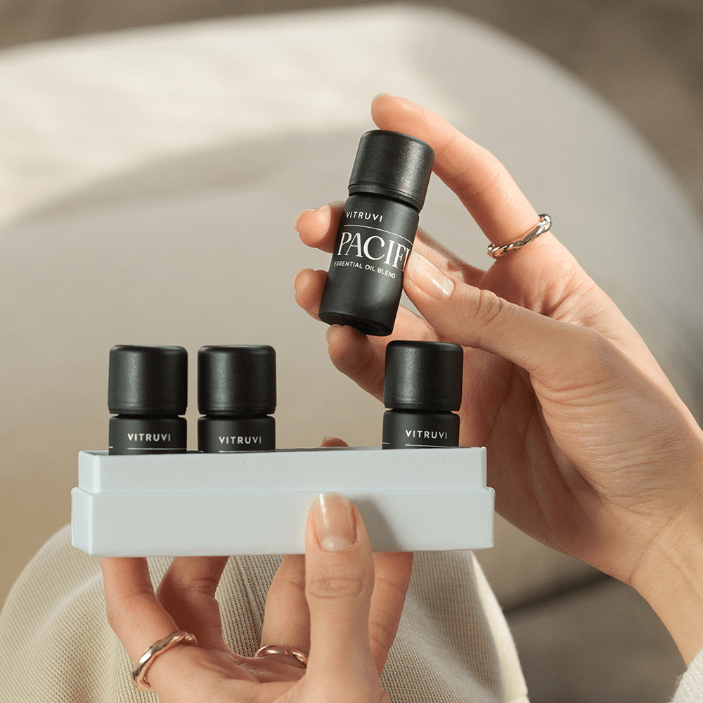 A person with neatly manicured nails is holding a small black bottle of Vitruvi Refresh Essential Oil Kit in one hand and a white tray with three more Vitruvi products in their other hand. The background is softly blurred, highlighting the elegance of these non-toxic home scents.