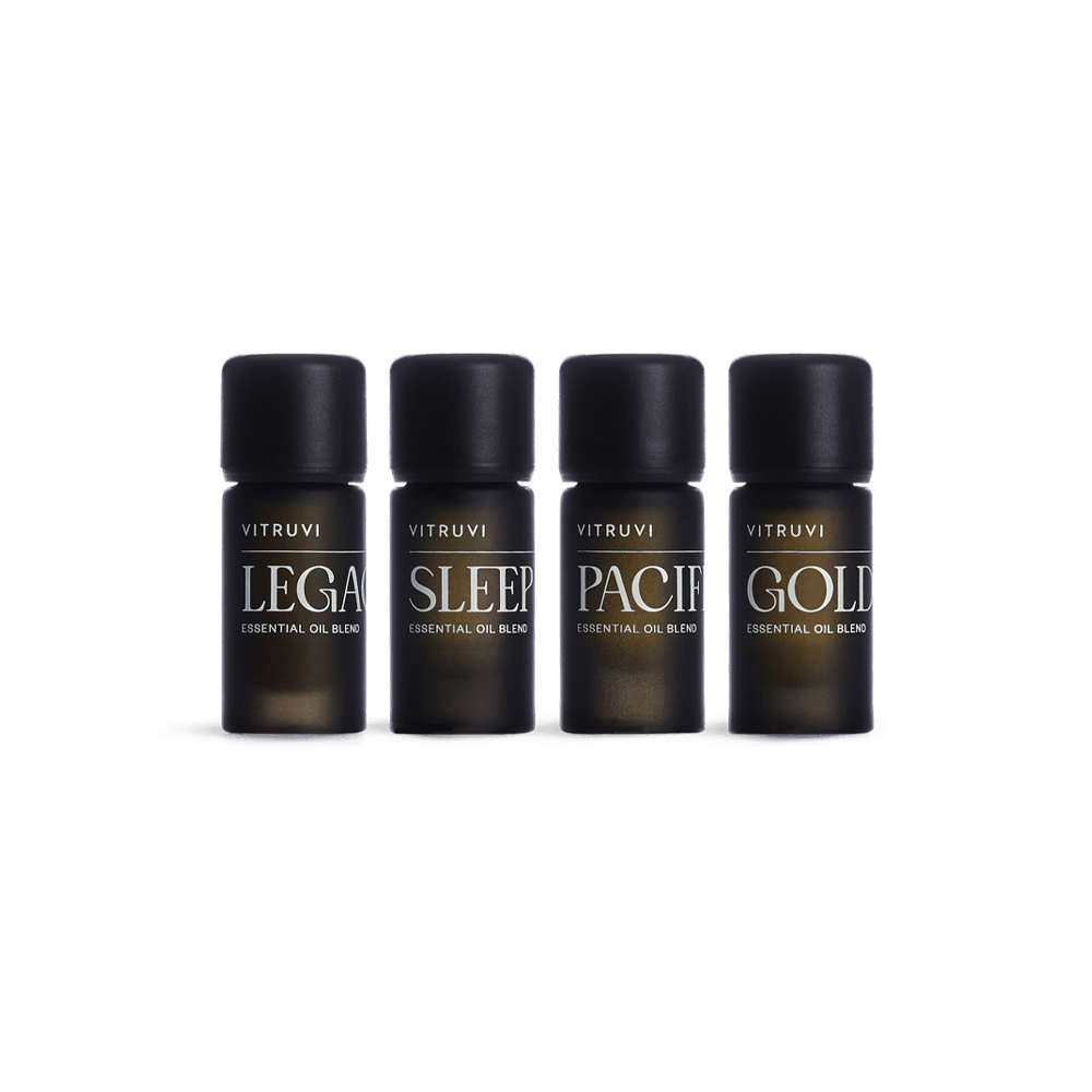 The Signature Essential Oil Kit by Vitruvi features a set of four black bottles, each labeled with its specific essential oil blend name: "Legacy," "Sleep," "Pacific," and "Golden." The soothing Sleep and grounding Legacy blends are prominently featured. The bottles are arranged in a straight line against a white background.