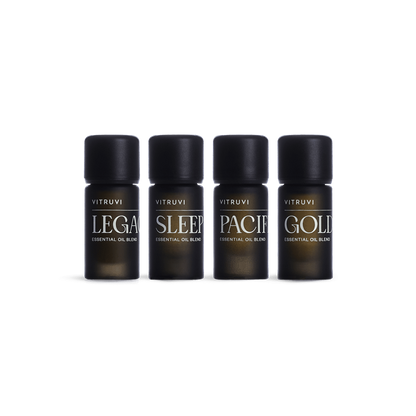 The Signature Essential Oil Kit by Vitruvi features a set of four black bottles, each labeled with its specific essential oil blend name: "Legacy," "Sleep," "Pacific," and "Golden." The soothing Sleep and grounding Legacy blends are prominently featured. The bottles are arranged in a straight line against a white background.