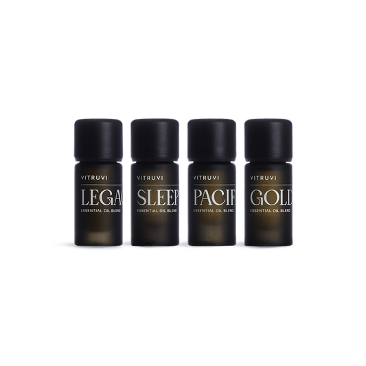 The Signature Essential Oil Kit by Vitruvi features a set of four black bottles, each labeled with its specific essential oil blend name: "Legacy," "Sleep," "Pacific," and "Golden." The soothing Sleep and grounding Legacy blends are prominently featured. The bottles are arranged in a straight line against a white background.