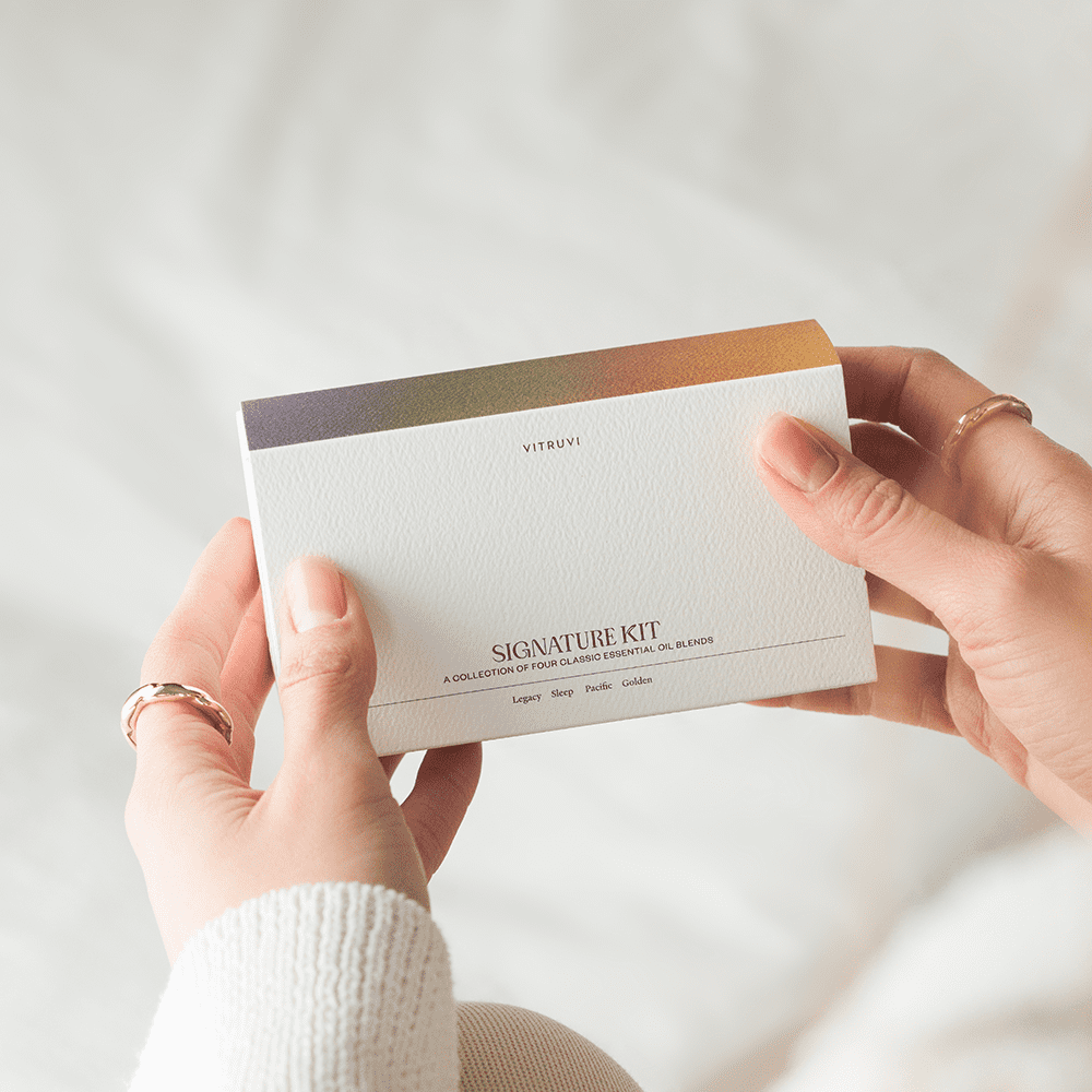 Close-up of hands holding a white box labeled "Signature Essential Oil Kit" by Vitruvi. The package is described as "A collection of four curated Vitruvi essential oil blends." The background is softly blurred, highlighting the focus on the product.