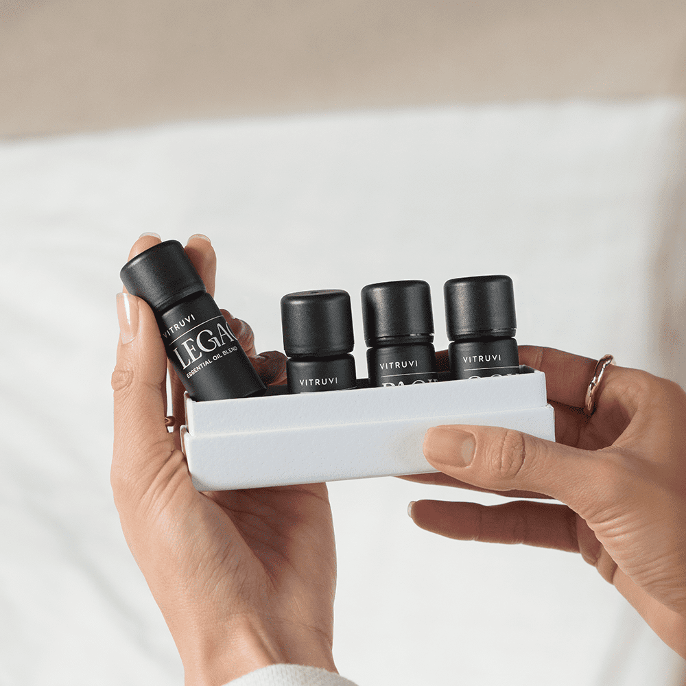 A person is holding the Vitruvi Signature Essential Oil Kit in a white container. The bottles are labeled with different names, and the individual is holding one bottle labeled "LEGACY" with both hands, showing neatly manicured nails. The background is blurred.