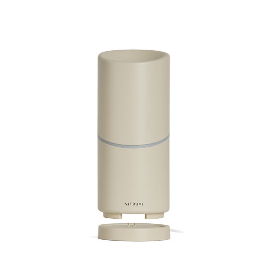 The Vitruvi Move Diffuser is a sleek, minimalist cylindrical diffuser with a smooth beige finish. It features a small, subtle logo near the base that reads "Vitruvi." This cordless diffuser sits on a matching round base and ensures you scent every corner with ease, no plug necessary.