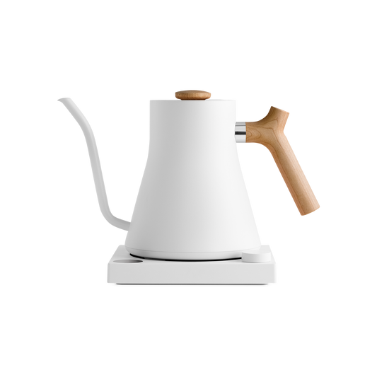 A modern white Fellow Wholesale Stagg EKG Electric Kettle with a matte finish is placed on a matching white base. It features a gooseneck spout and light wooden handle and lid knob. The sleek design combines variable temperature control with aesthetics, making it ideal for precision pouring in pour-over coffee.
