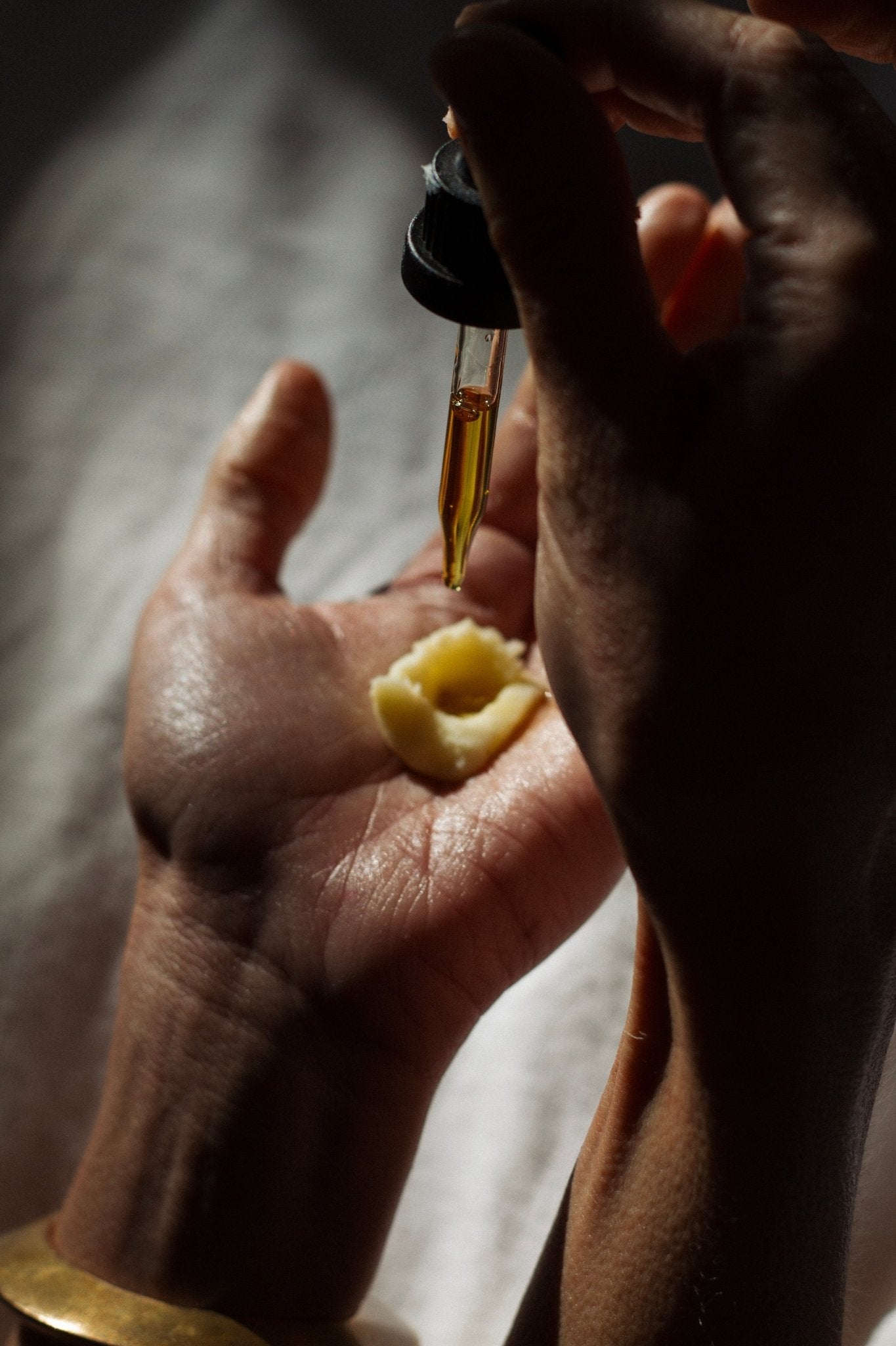 A close-up of two hands, one holding a small scoop of cream and the other dispensing Emanations Nourishing Face and Hair Oil-Animal-based from Summer Solace Tallow using a dropper. The background is softly lit with a neutral tone, adding to the luxury skincare ambiance. A gold bracelet gleams on the wrist holding the cream.