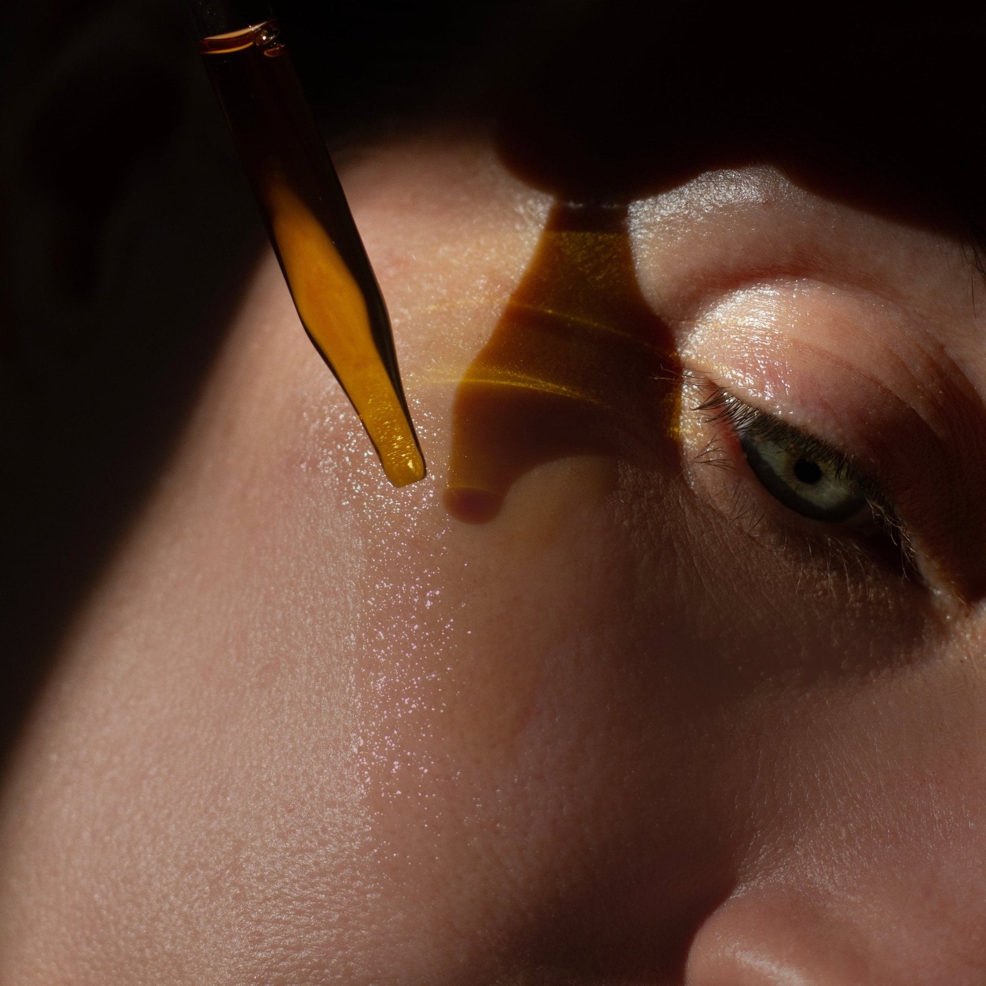 Close-up shot of an eye with pale skin, illuminated by light. A dropper releases a droplet of Summer Solace Tallow's Emanations Nourishing Face and Hair Oil-Animal-based onto the skin just below the eye, highlighting the texture and glistening effect. The scene conveys a sense of luxury skincare routine and facial care.