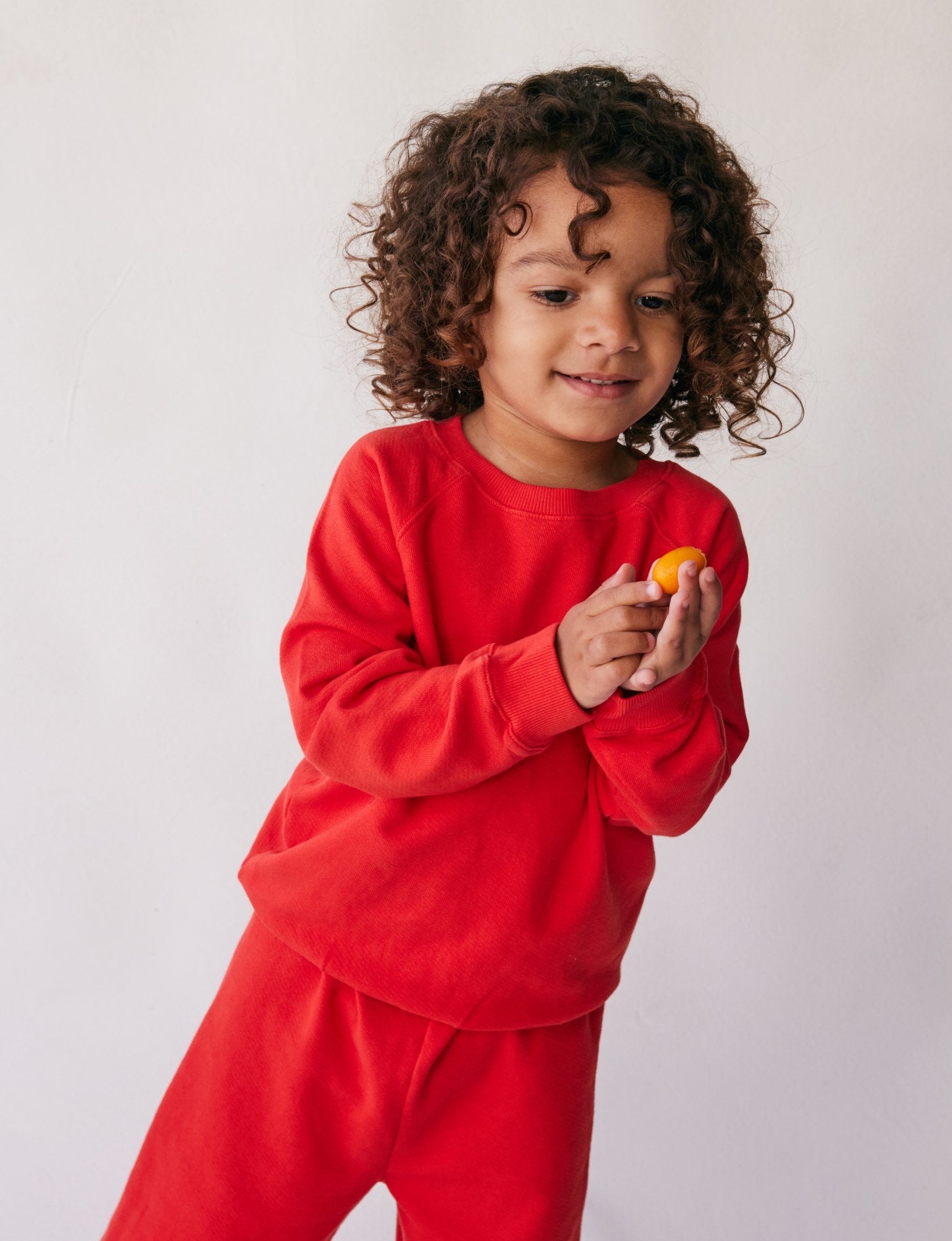 A young child with curly hair, dressed in The Lil' Classic Crewneck by all the babies along with matching pants, smiles while looking at a small yellow object held in their hands. The background is plain white.