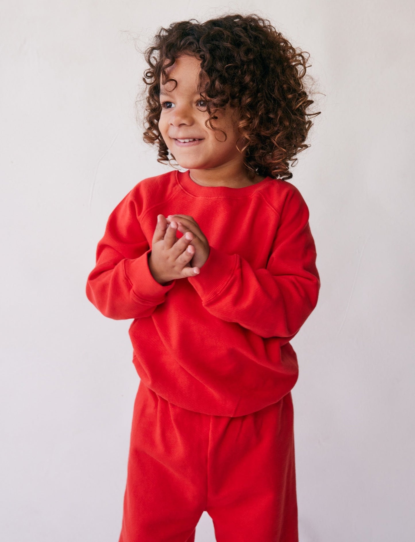 A young child with curly hair smiles while looking to the side, wearing The Lil' Classic Crewneck in bright red along with matching pants by all the babies, set against a plain, light-colored background. The child's hands are clasped together in front of them.