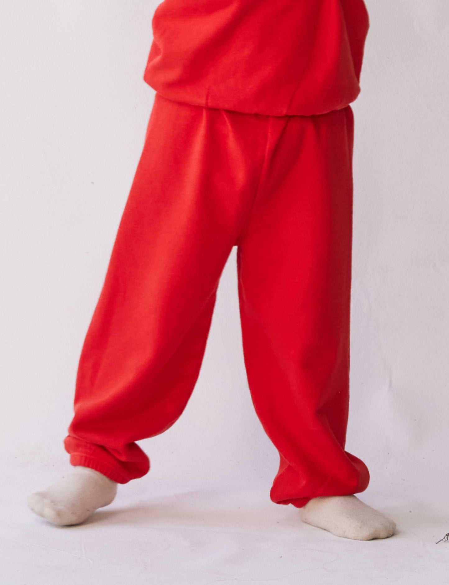 A person is wearing The Lil' Classic Sweatpant in bright red from all the babies, paired with a matching red shirt crafted from organic French terry cloth. White socks are visible at the bottom, against a plain white background. Only the lower half of the body is shown.