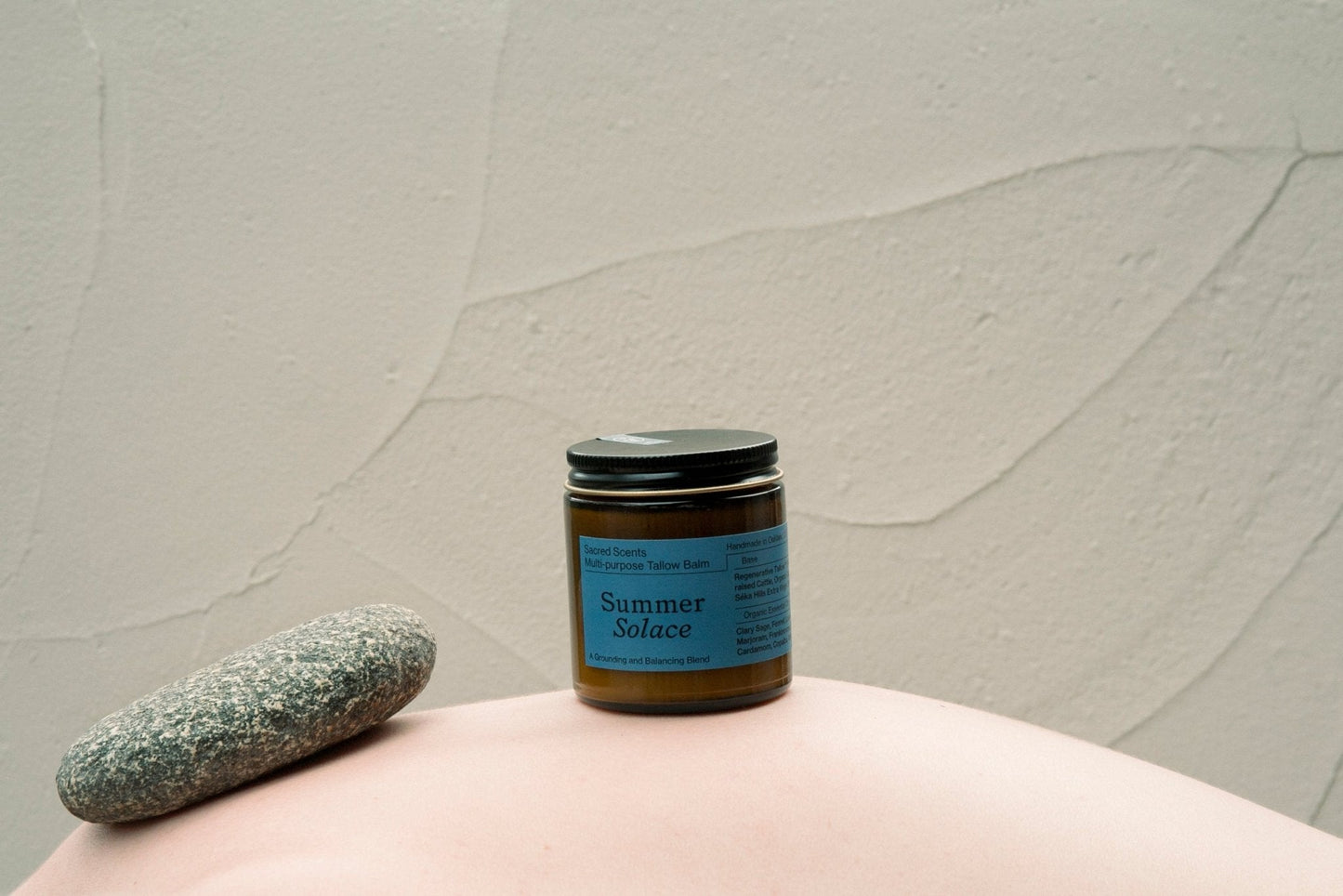 A jar labeled "Sacred Scents Spiritual Balm - Regenerative Tallow®" by Summer Solace Tallow sits on a smooth surface beside a gray stone, hinting at its herbal tallow balm infused with anti-inflammatory essential oil. The background is a light, textured wall.