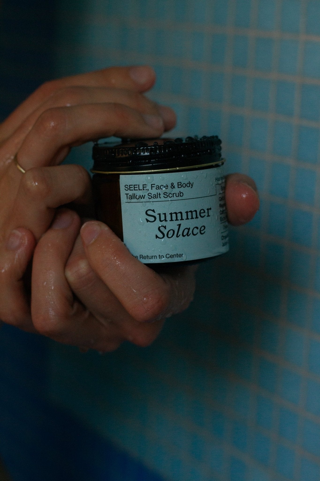 Hands holding a brown jar labeled "SEELE Celtic Sea Salt® Scrub - Regenerative Tallow®" from Summer Solace Tallow, an exfoliating scrub with a marine-like scent, set against a background of blue tiled wall.