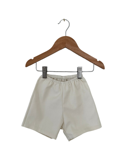 A pair of beige Parc Shorts in Hemp Twill from cabane childrenswear, featuring an elastic waistband, hangs on a wooden hanger against a black background.