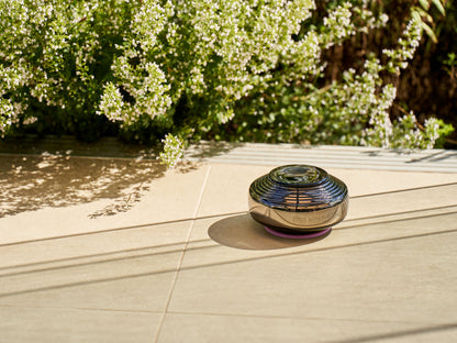 A small, disc-shaped speaker emits soft music while placed on Somavedic USA's Anthracite-tiled sunny patio. Greenery in the background casts shadows on the tiles, creating a peaceful outdoor atmosphere.