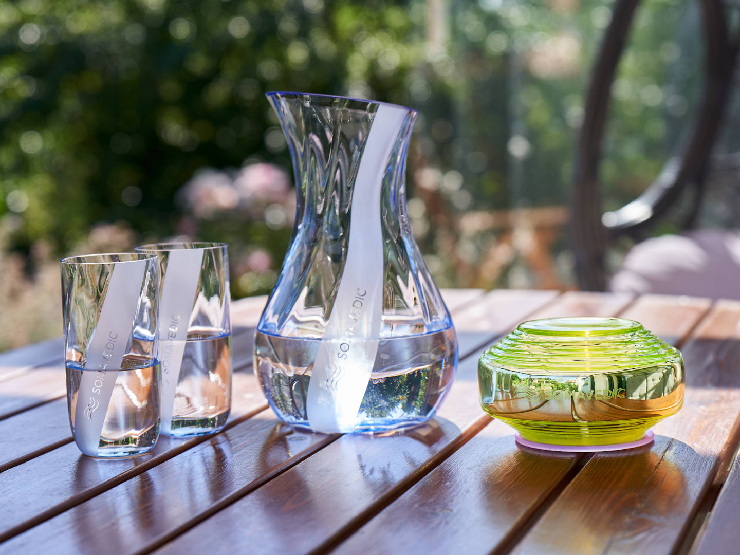 A clear glass carafe filled with water sits on a wooden table alongside two matching clear glasses, featuring the innovative Somavedic USA Vedic for water structuring to reduce oxidative stress. Next to them is a round, decorative green and yellow glass candle holder. The background shows a blurred outdoor garden scene.