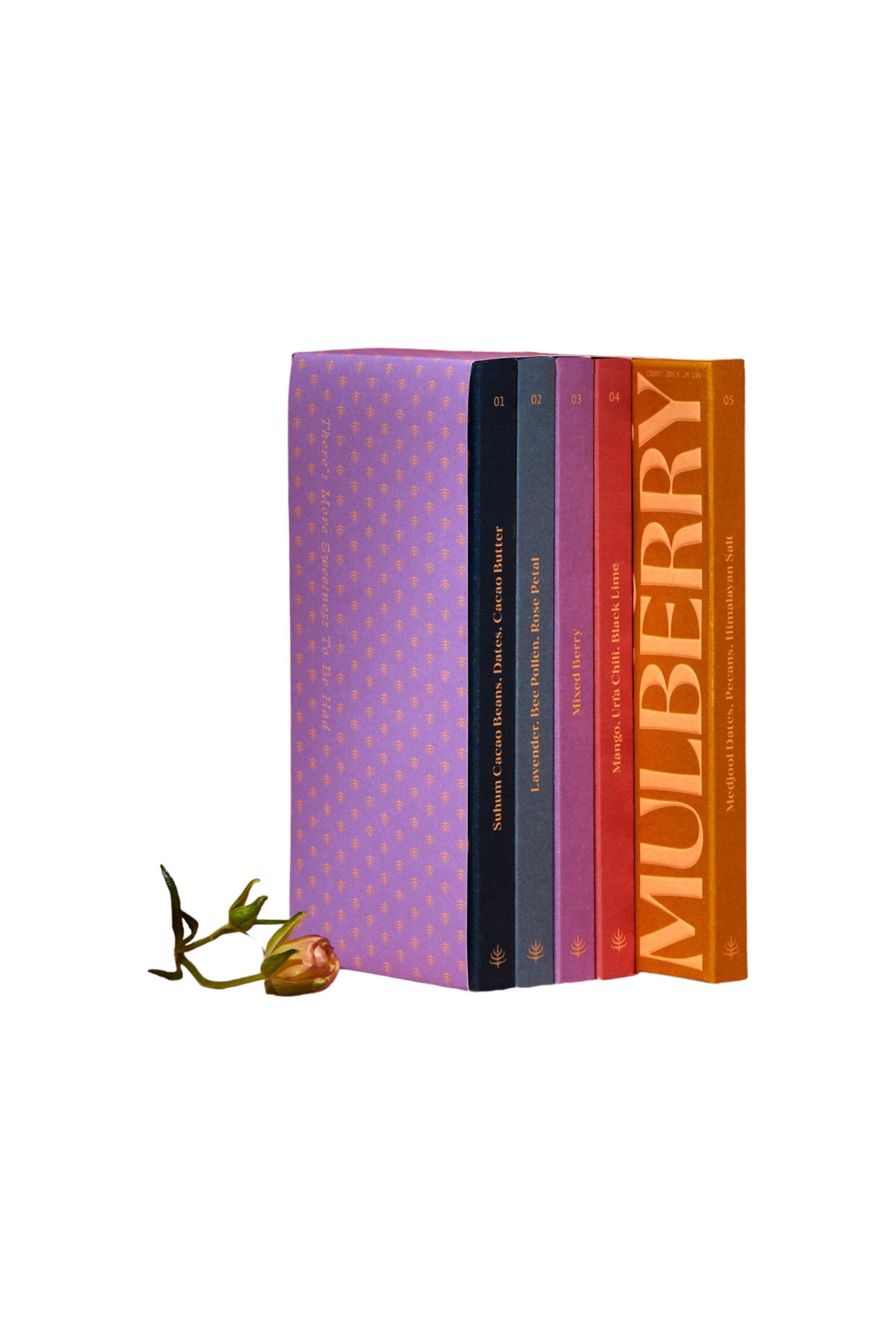 A set of five "Discovery Collection" books by Spring & Mulberry, each with a colorful cover and labeled from 01 to 05 in gold text on the spine, stands upright against a black background. At the base of the books lies a single, small rosebud with its stem extending slightly to the left, evoking the rich simplicity of date-sweetened chocolate.