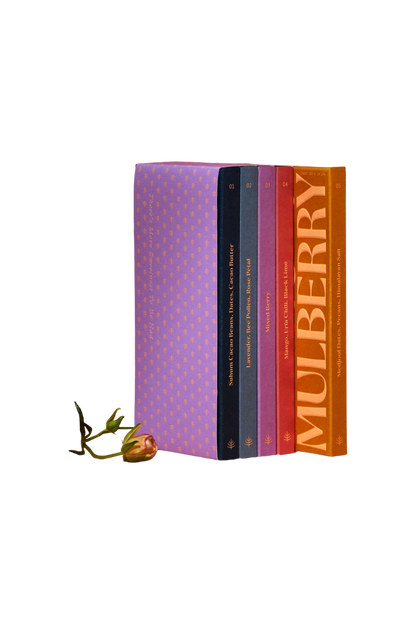 A set of five "Discovery Collection" books by Spring & Mulberry, each with a colorful cover and labeled from 01 to 05 in gold text on the spine, stands upright against a black background. At the base of the books lies a single, small rosebud with its stem extending slightly to the left, evoking the rich simplicity of date-sweetened chocolate.