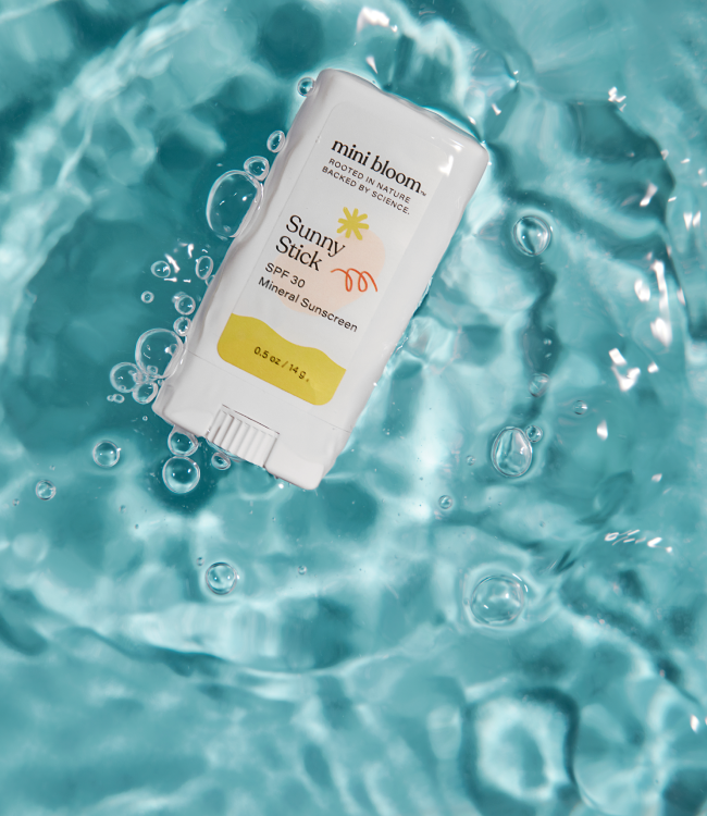 A white rectangular sunscreen stick with rounded edges and a twist-up base labeled "Sunny Stick Mineral Sunscreen" by mini bloom floats in clear, aqua-colored water surrounded by small bubbles. The product is designed as a baby sunscreen.