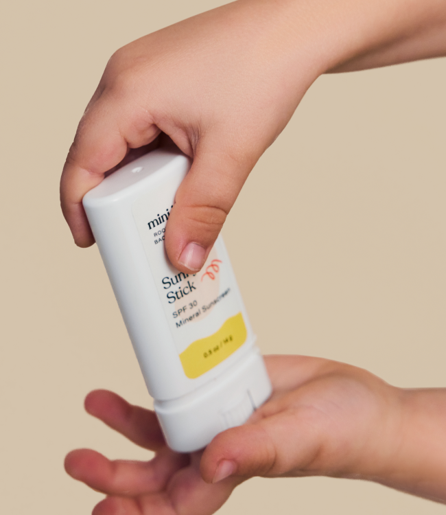 A close-up of a person holding the Sunny Stick Mineral Sunscreen by mini bloom with their right hand, applying it to the palm of their left hand. The sunscreen stick features a white container with yellow and white labeling, perfect for gentle application.