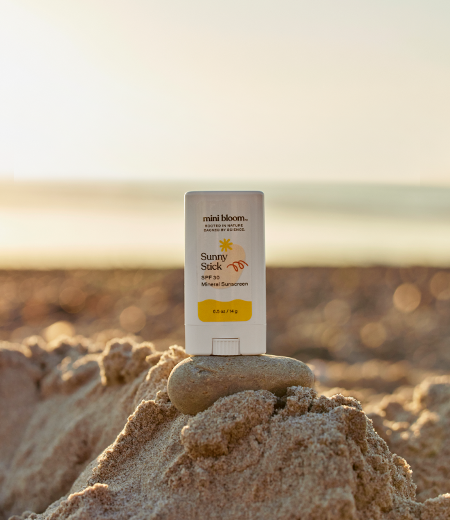 A Sunny Stick Mineral Sunscreen by mini bloom rests on a small sand mound with a smooth stone beneath it. The background features a blurred, sunlit beach and calm waters in the distance, making it perfect for gentle baby sunscreen protection.