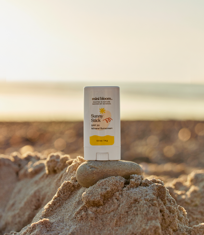 A Sunny Stick Mineral Sunscreen by mini bloom rests on a small sand mound with a smooth stone beneath it. The background features a blurred, sunlit beach and calm waters in the distance, making it perfect for gentle baby sunscreen protection.