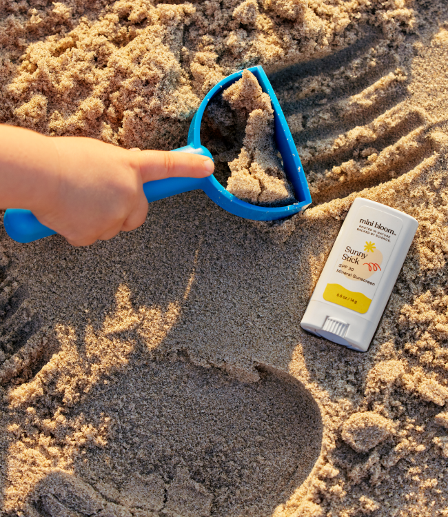 A child's hand holding a blue sand scoop, digging in the sandy beach. Next to the scoop is a stick of baby sunscreen labeled "Sunny Stick Mineral Sunscreen" by mini bloom, resting on the sand. Sunlight casts shadows, highlighting the textures of the sand and the products.