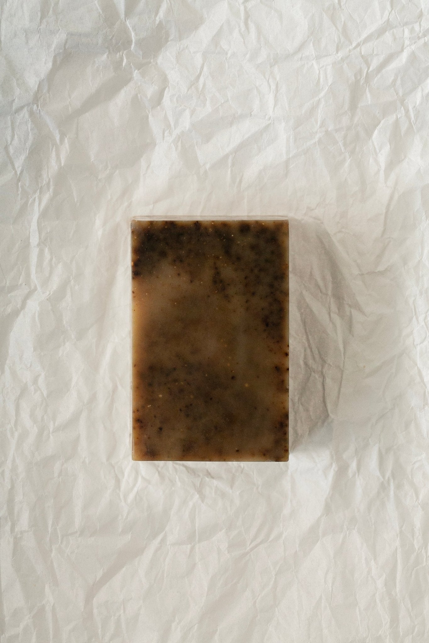 A rectangular bar of Third Wave Coffee Bar Soap - Regenerative Tallow™ by Summer Solace Tallow, featuring a mottled brown appearance, is displayed on white crumpled paper. The handmade soap exhibits visible specks throughout its surface, indicating it may contain natural ingredients or exfoliants from Northern California pasture-raised cattle.