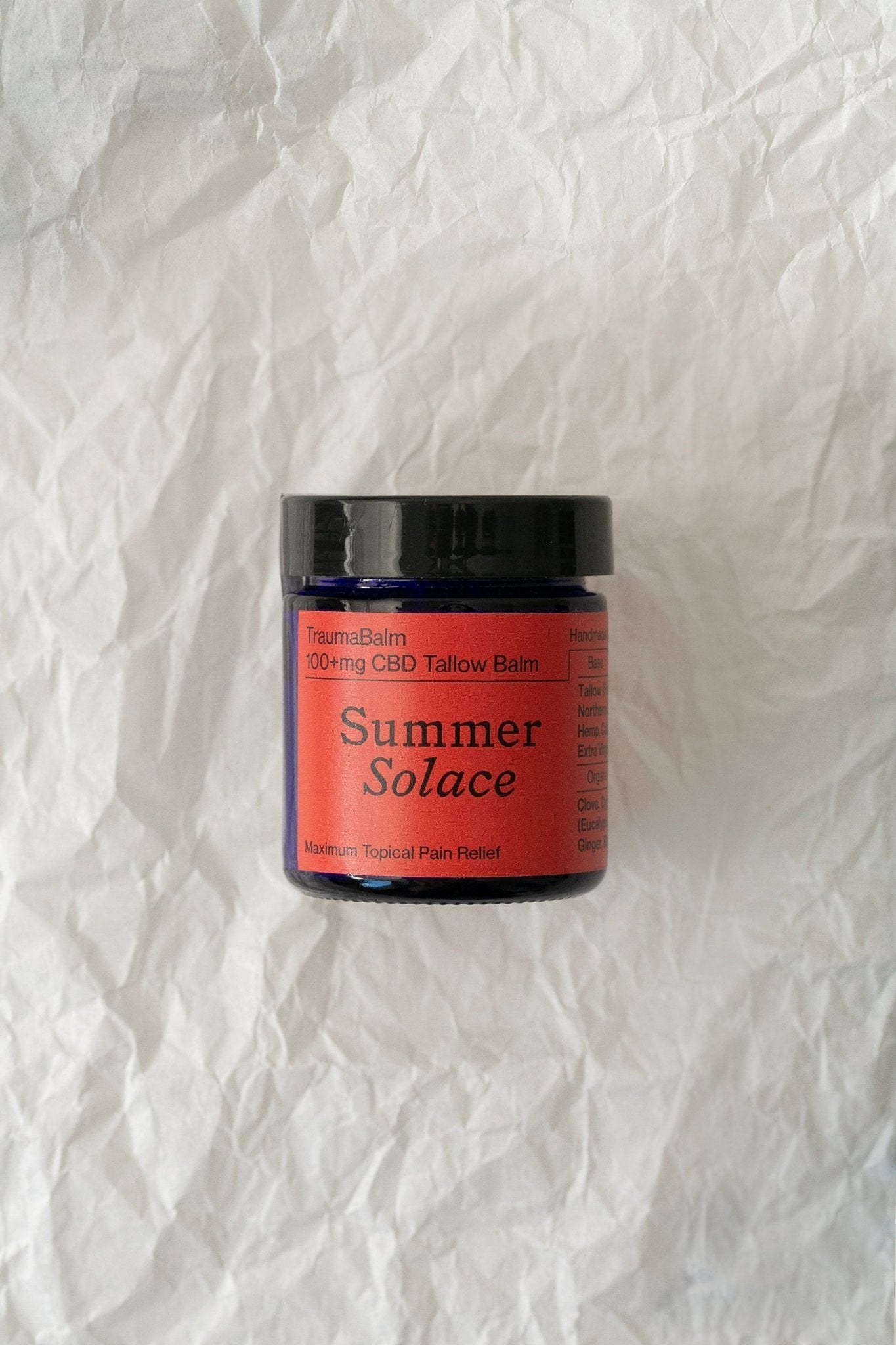 A black jar labeled "TraumaBalm Soothe Soreness - Regenerative Tallow™" by Summer Solace Tallow, known for its anti-inflammatory benefits and maximum topical pain relief, is placed on a crumpled white paper background. The product label is vivid red with black and white text.