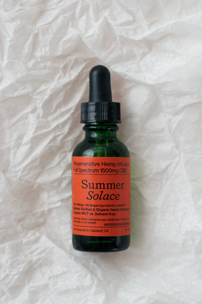 A small green glass bottle with a black dropper cap lies on crumpled white paper. The label reads "Summer Solace Tallow" and indicates it contains TraumaBalm Soothe Soreness - Regenerative Tallow™, a regenerative hemp infusion of full-spectrum 1500mg CBD. The text mentions organic ingredients and being hand-poured in Oakland, CA.