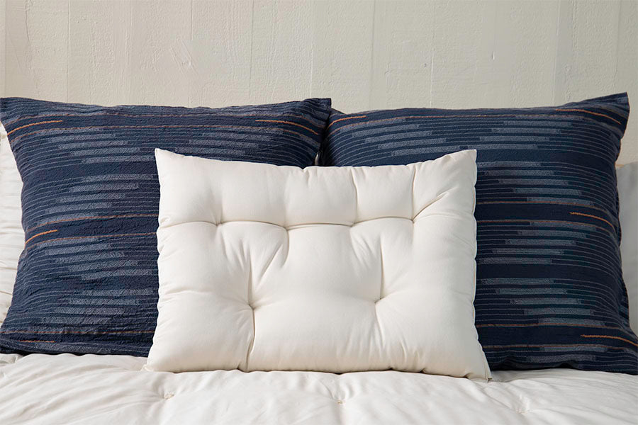 Three pillows are arranged on a bed. Two large, dark blue pillows featuring horizontal stripes sit at the back, with a smaller tufted white pillow positioned in front. The white bedding is accented by a Shepherd's Dream Wool Contour Sleep Pillow for enhanced neck support, set against a light-colored wall.