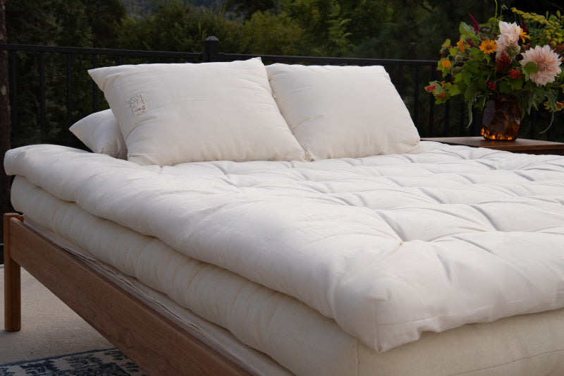 A neatly arranged bed featuring the Shepherd's Dream Snuggle Mate Wool Topper, complete with fluffy white pillows, is set on a wooden frame outside. A vase filled with vibrant flowers sits on the side table, while a black railing adorned with greenery serves as the backdrop.