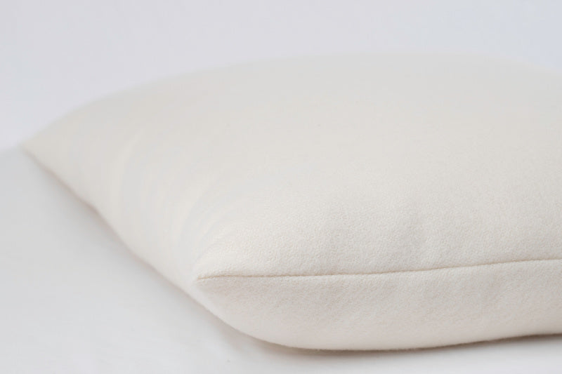 A close-up image of the allergen-reducing, off-white All Wool Dream Pillow from Shepherd's Dream, resting on a similarly colored surface. The pillow appears soft and slightly textured, with a smooth seam along its edge. The simple background accentuates the pillow's material and design.