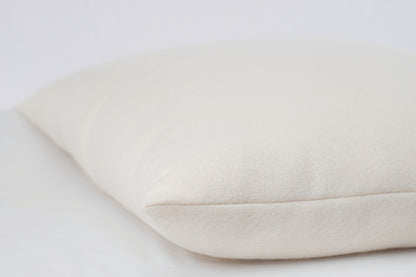 A close-up image of the allergen-reducing, off-white All Wool Dream Pillow from Shepherd's Dream, resting on a similarly colored surface. The pillow appears soft and slightly textured, with a smooth seam along its edge. The simple background accentuates the pillow's material and design.