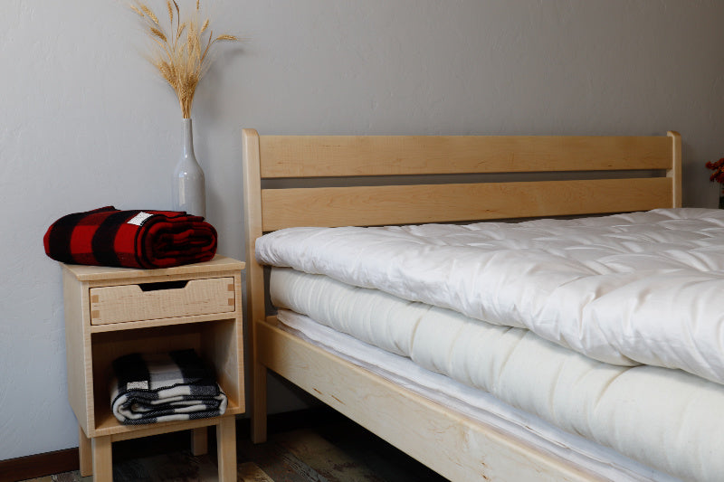 A neatly made bed with a white comforter sits alongside a wooden nightstand, featuring a folded red and black blanket. The All Wool Mattress by Shepherd's Dream promises supportive sleep, while a vase with dried plants adds a decorative touch.