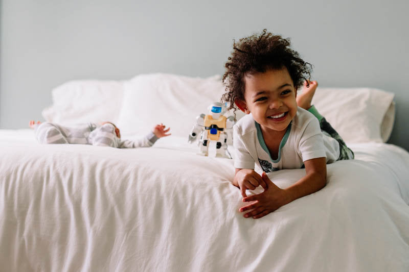 A child smiles while lying on a Shepherd's Dream All Wool Mattress, with a toy robot next to them. Another child is in the background, partially covered by EcoWool® blankets. The room is softly lit, creating a warm and cozy atmosphere perfect for a peaceful night’s sleep.