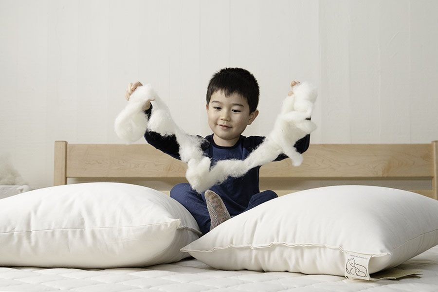 A young child sits on a bed crafted from a Cascade Latex Mattress by Shepherd's Dream, nestled between two large pillows, holding a fluffy piece of white cotton. The room boasts a simple and minimalist design with light-colored walls and bedding.