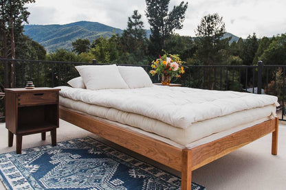 A bed featuring the Snuggle Mate Wool Topper by Shepherd's Dream, styled with white organic cotton sateen bedding and pillows, is situated on a rug on an outdoor patio framed by stunning mountain views. A small wooden side table displays a stack of items, while a vase filled with vibrant flowers enhances the decor. In the background, trees and mountains create a picturesque setting.