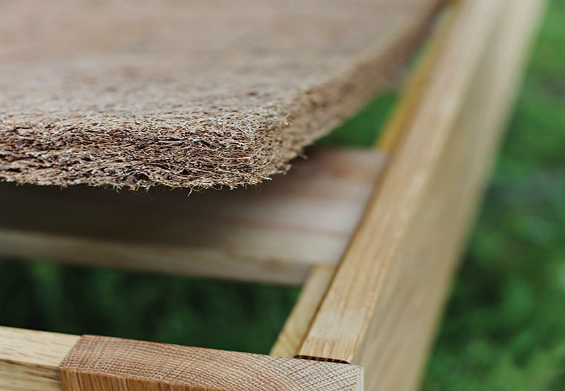 A close-up of a wooden frame features the textured CocoCoir Comfort Pad by Shepherd's Dream, with its fiber-like coconut coir surface visible. The blurred green background hints at an outdoor setting.