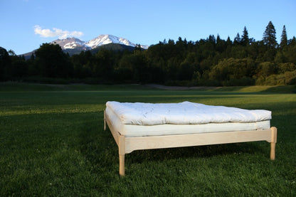 The Ashland Organic Wool Topper by Shepherd's Dream is showcased on a bed with a light wooden frame and white mattress, elegantly positioned outdoors on a grassy field. Green trees and a snow-capped mountain rise in the background beneath a clear blue sky, highlighting the natural breathability of this tranquil scene.