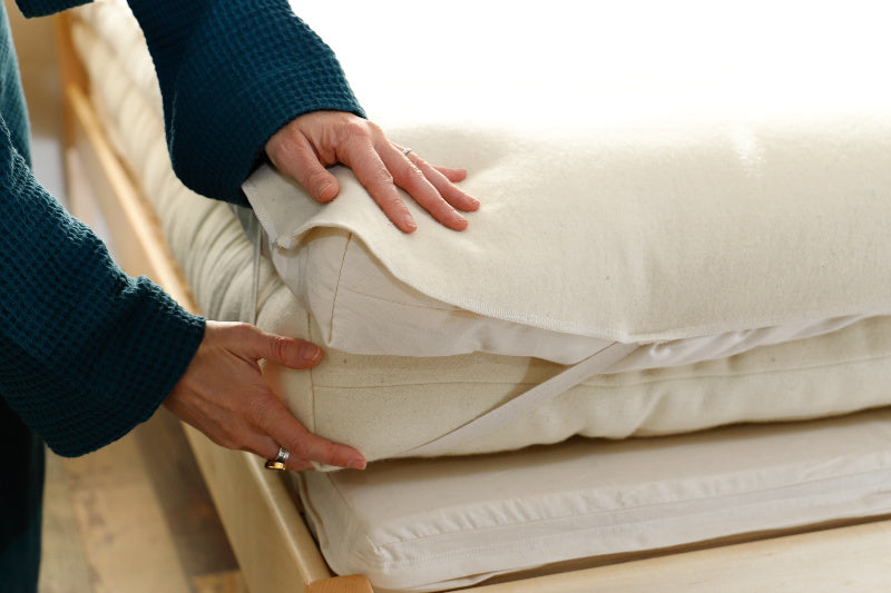 A person wearing a dark, textured sweater adjusts a white mattress cover with elastic straps, revealing the Shepherd's Dream Wool Mattress Protector underneath. The well-lit room highlights the clean and neat bedding arrangement.