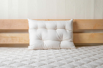 A single white, tufted pillow rests against a light wood headboard on the Shepherd's Dream Cascade Latex Mattress, covered with an organic cotton encasement. The off-white wall behind creates a serene and minimalist bedroom setting.