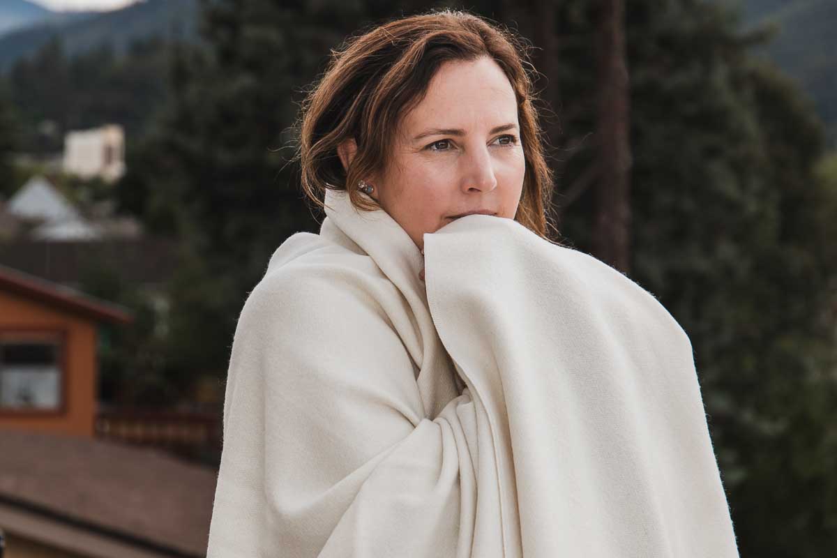 A woman wrapped in a Shepherd's Dream Organic Merino Wool Blanket stands outdoors, gazing thoughtfully into the distance, with forested hills and a house visible in the background.