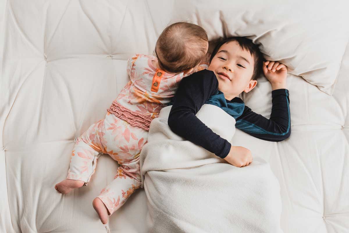 A young child sleeps on a bed, cuddling with a baby atop the Snuggle Mate Wool Topper by Shepherd's Dream. Covered with a light blanket, the baby wears floral pajamas while the older child dons a blue and black outfit. The cozy scene of warmth and affection is enhanced by the softness of organic cotton sateen.