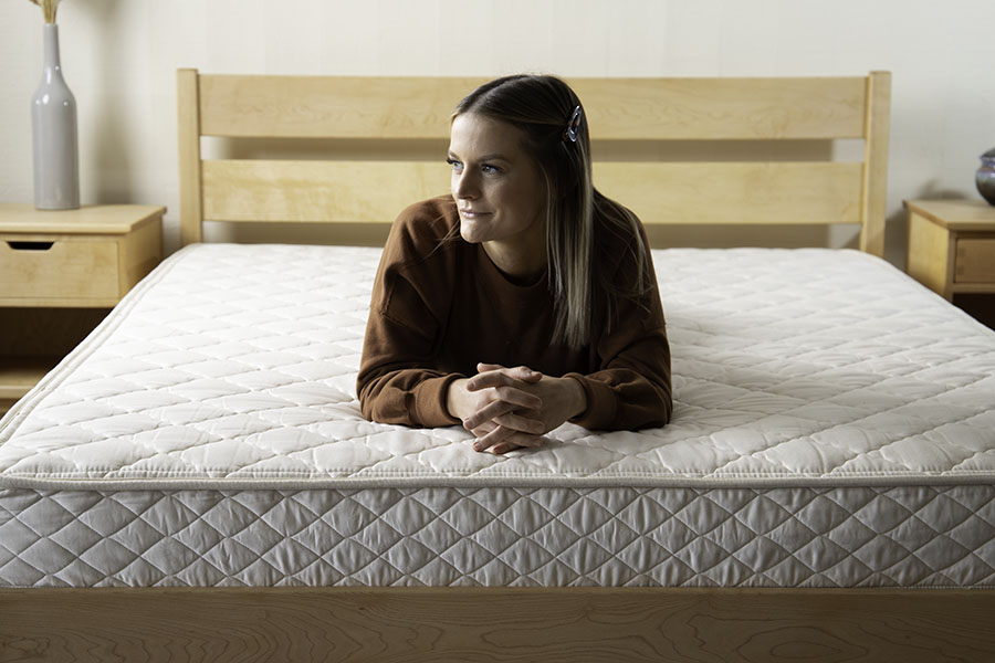 A woman with long hair secured by a clip sits on a large bed featuring the Cascade Latex Mattress from Shepherd's Dream. She is wearing a brown long-sleeve top and looking to the side. The bedroom decor, including a wooden bed frame and bedside tables, enhances the serene ambiance.