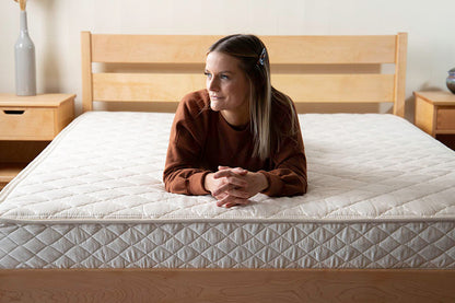 A woman with long hair secured by a clip sits on a large bed featuring the Cascade Latex Mattress from Shepherd's Dream. She is wearing a brown long-sleeve top and looking to the side. The bedroom decor, including a wooden bed frame and bedside tables, enhances the serene ambiance.