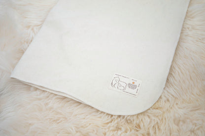 A luxurious, cream-colored blanket crafted from natural wool lies on a fluffy white backdrop, featuring a small label adorned with minimalist designs and text showcasing a simple line drawing of two cats. Ideal as baby bedding, this is the Washable Wool Crib Mattress Protector by Shepherd's Dream.