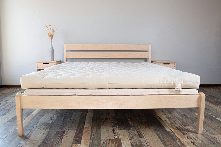 A minimalist wooden bed with a thick All Wool Mattress from Shepherd's Dream is centered in a room with wooden flooring. Two small wooden nightstands flank the bed, and a tall vase with dried wheat decorates one nightstand. The walls are plain and light-colored.