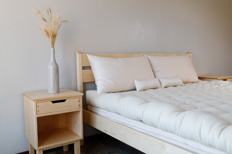 A minimalist bedroom showcases a light wood bedframe paired with a nightstand. The All Wool Mattress by Shepherd's Dream is topped with a white comforter and pillows. The nightstand, featuring a single drawer, is elegantly decorated with a vase of dried wheat stalks. Soft beige walls enhance the serene atmosphere.