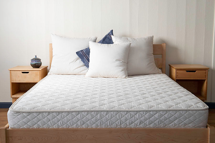 A neatly made bed with white sheets and four pillows rests on a Shepherd's Dream Cascade Latex Mattress, perfectly positioned between two wooden nightstands. Each nightstand has an open shelf and a drawer. A small decorative pot is placed on one of the nightstands, adding charm to the serene setup.