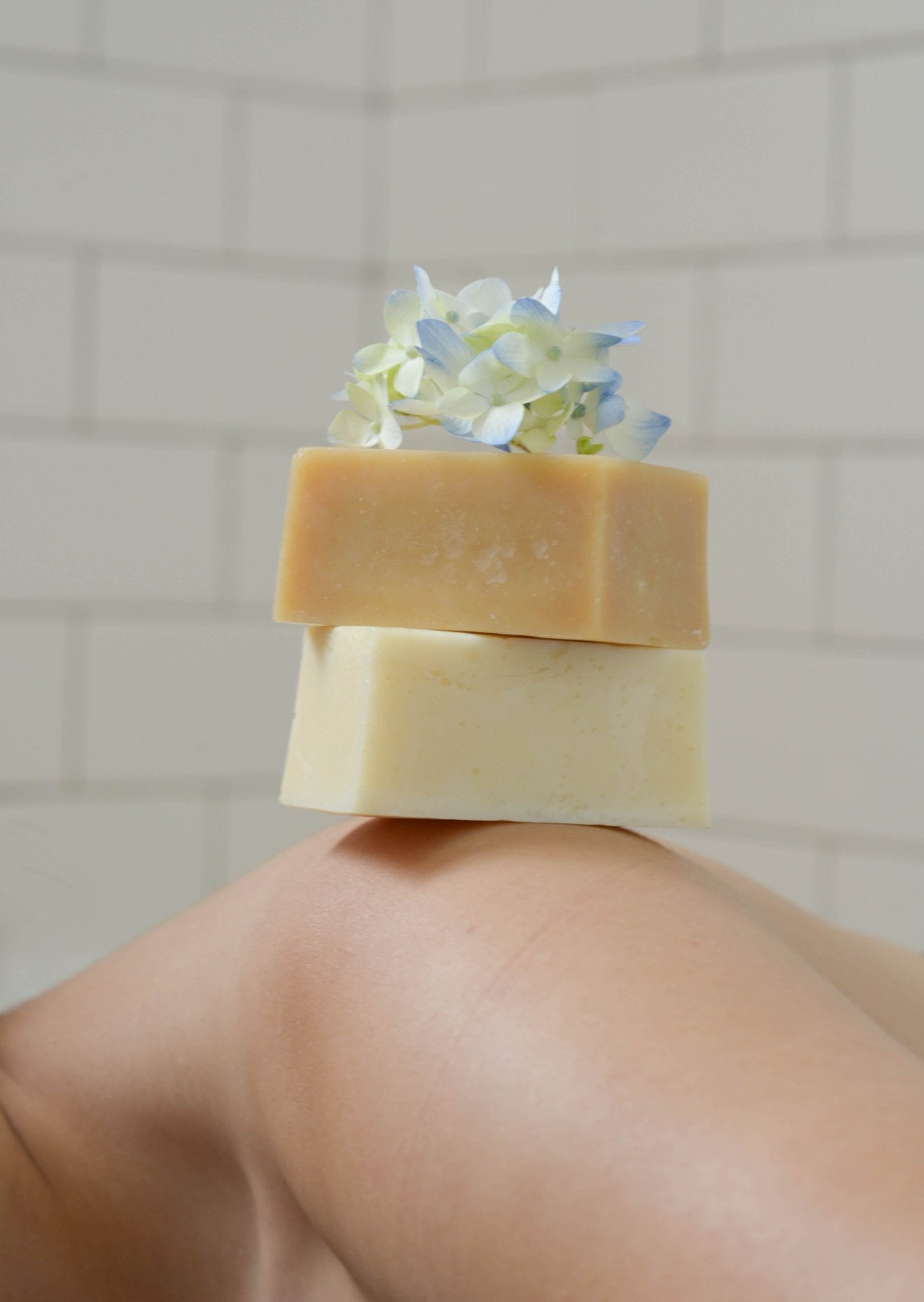 Close-up of two stacked Summer Solace Tallow Violet and Lemon Basil A2/A2 Milk Soaps - Regenerative Tallow® on a person's shoulder. The top soap bar has a light brown hue, while the bottom one is creamy beige. A small cluster of pastel blue and white flowers rests atop the soap bars. The background features white tiled walls.