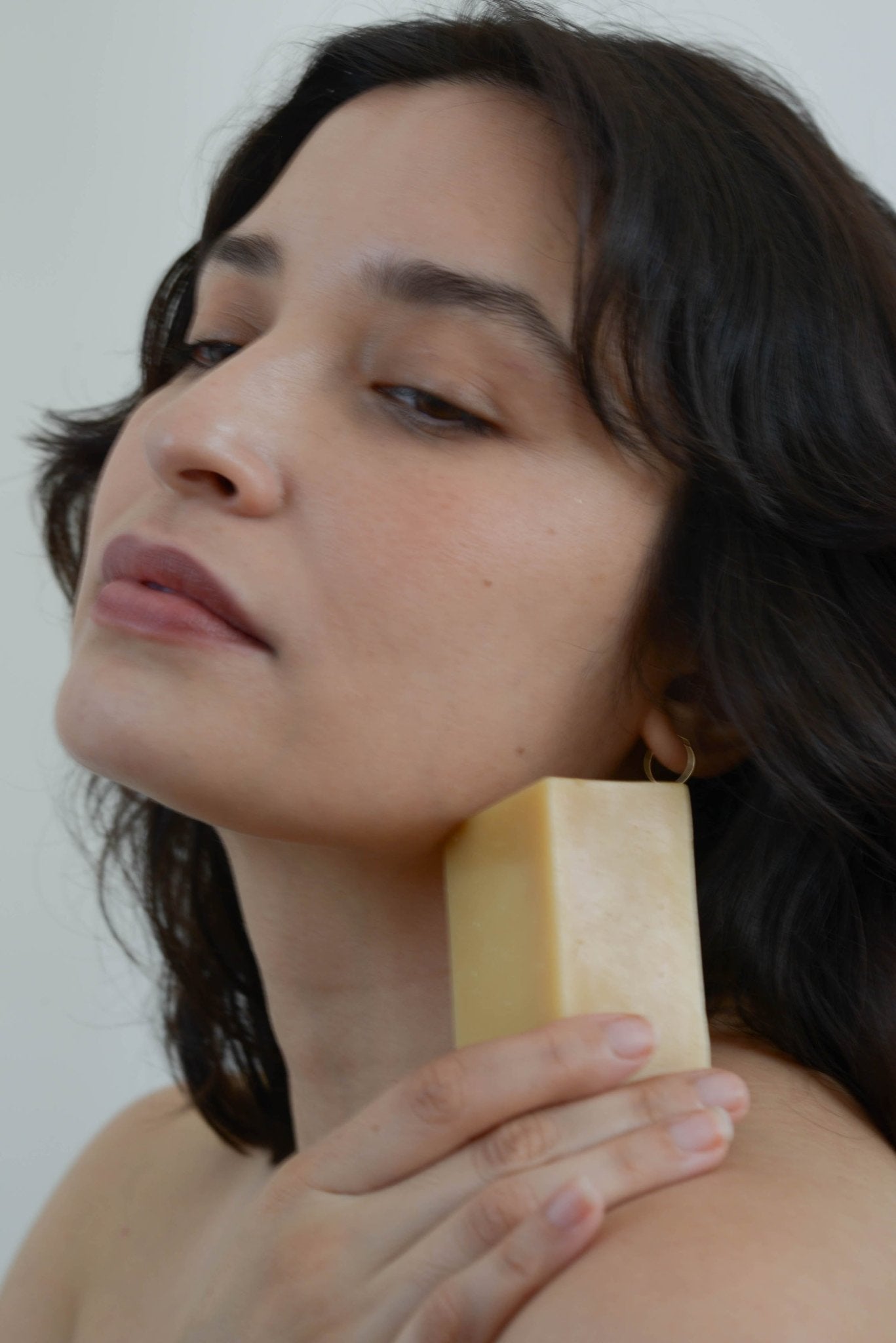 A person with long dark hair gently pressing a bar of Summer Solace Tallow's Violet and Lemon Basil A2/A2 Milk Soap - Regenerative Tallow® against their jawline. Their eyes are closed, and they have a calm expression. The background is plain and light-colored.